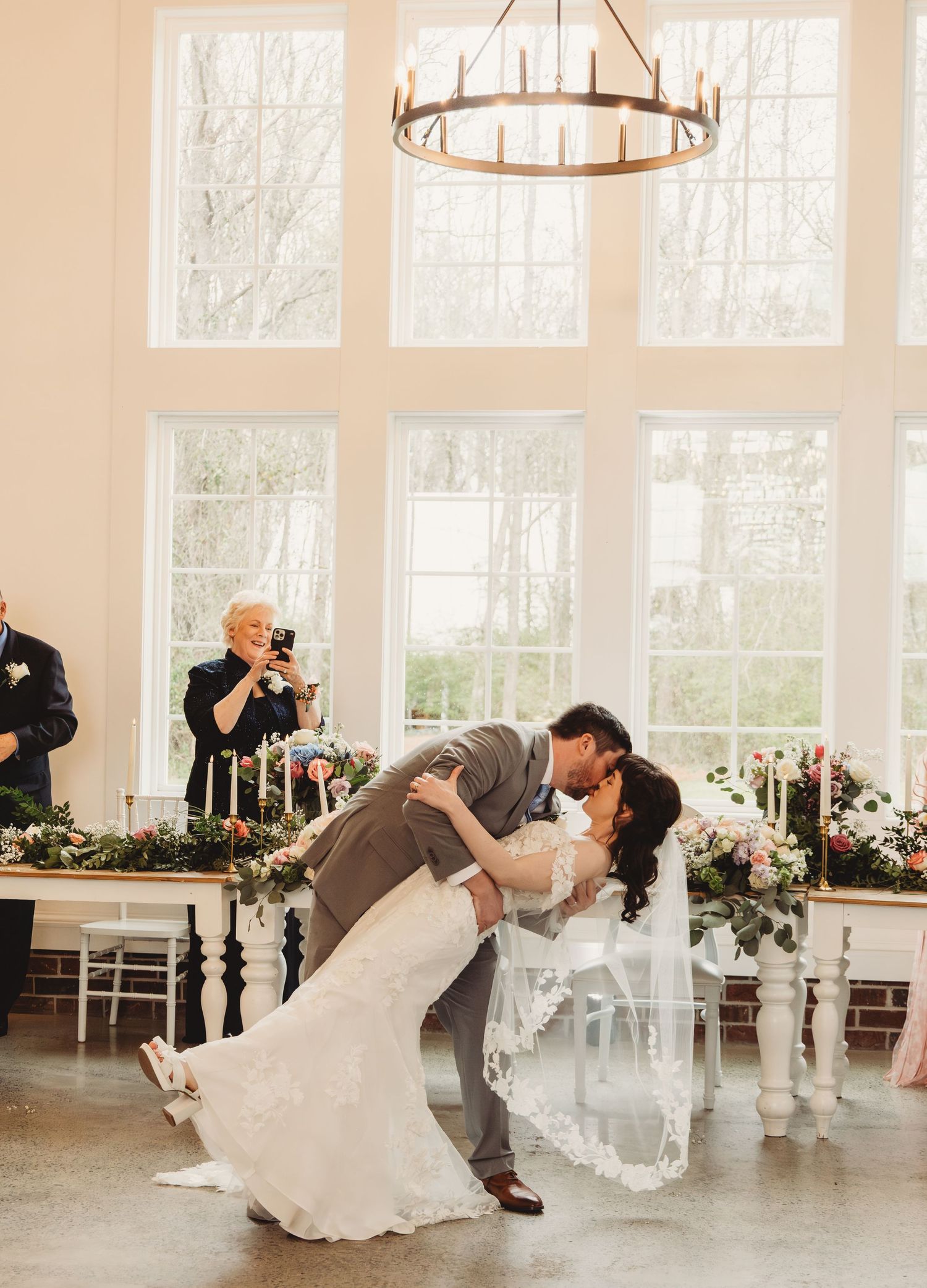 Bride and groom share a romantic dip kiss at the altar in a bright white wedding venue with floral arrangements.