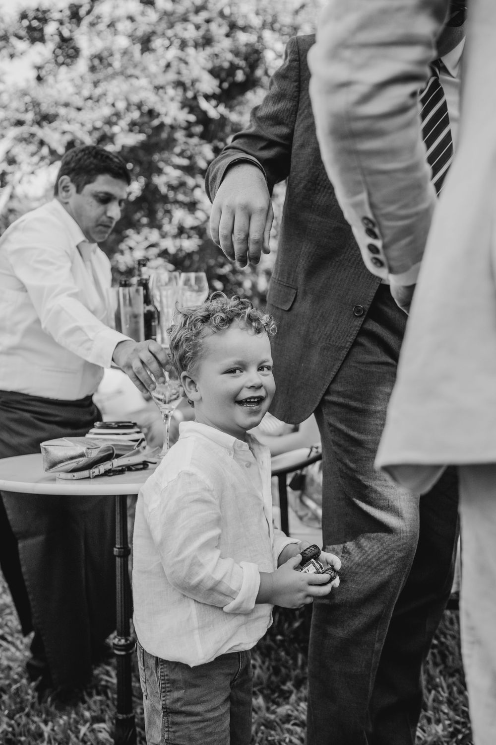 Black and white photo of a joyful young boy laughing at an outdoor wedding reception with suited men nearby.