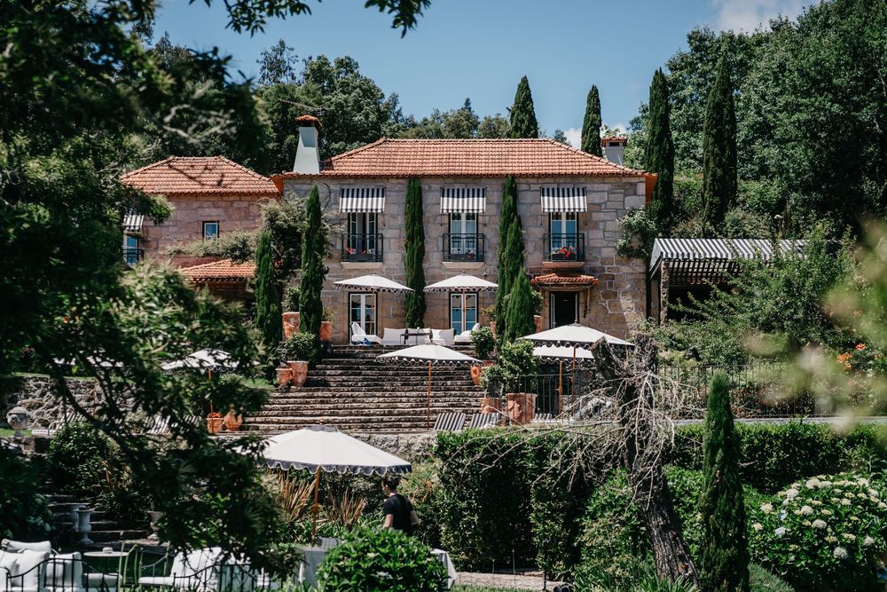 Stone villa with terracotta roof surrounded by lush cypress trees and manicured gardens with white umbrella dining terrace.