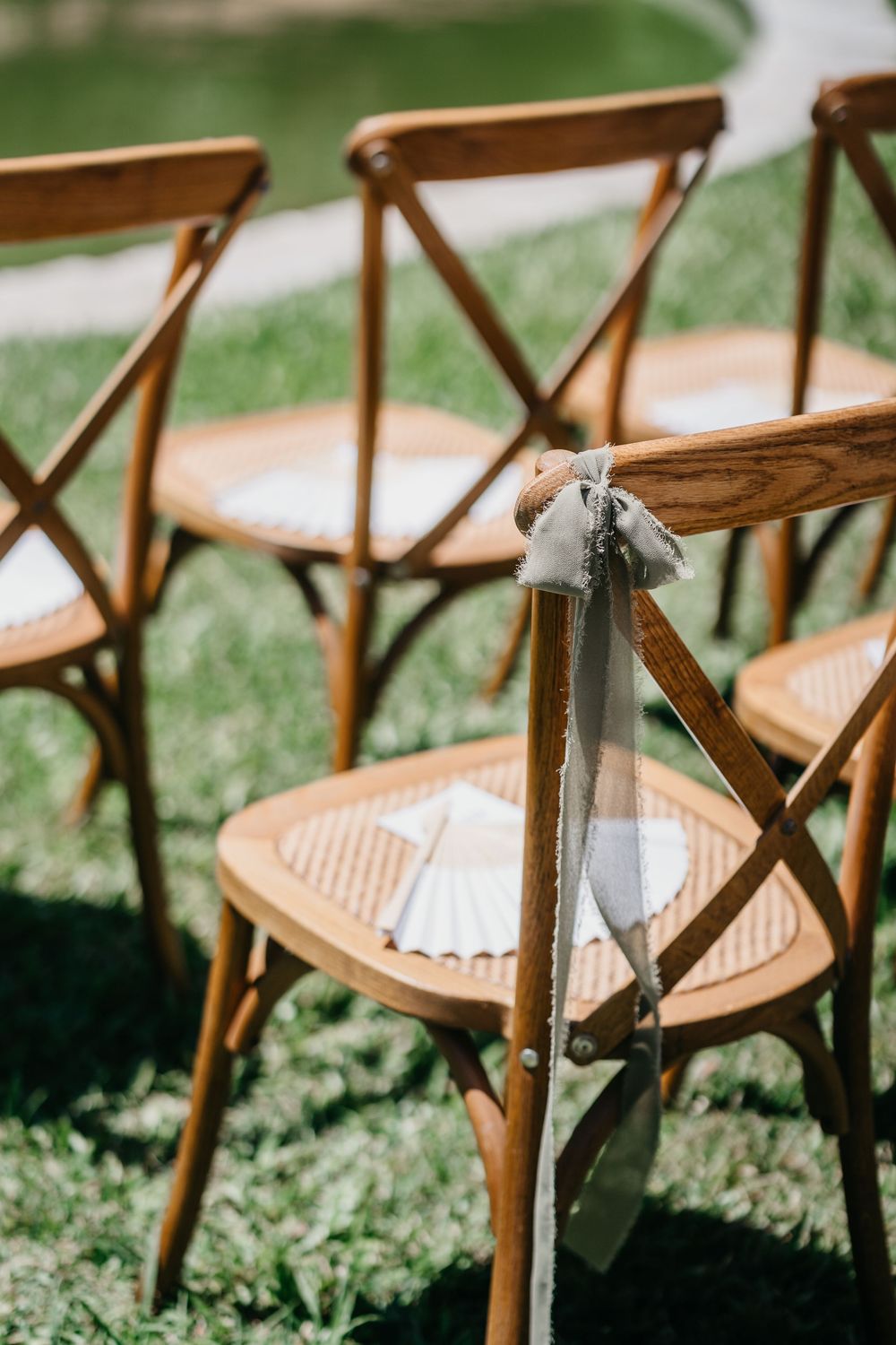 Wooden cross-back chairs with ribbon decor on green grass at an outdoor wedding ceremony.