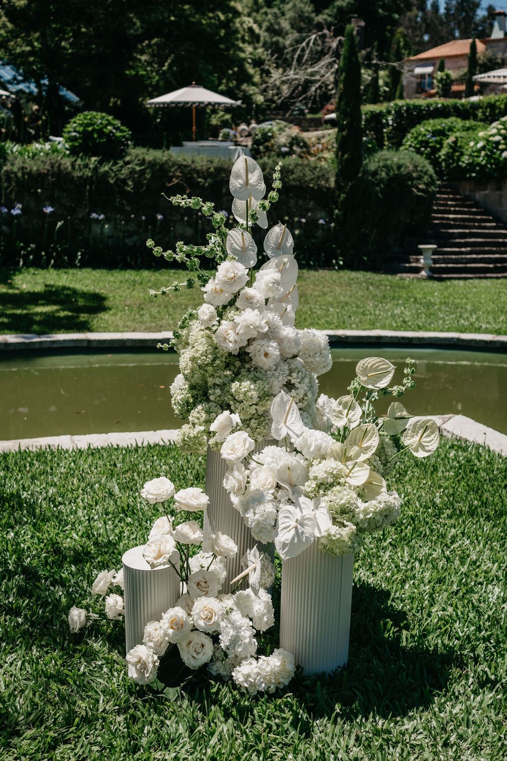 Elegant white floral arrangement with roses and anthuriums on ribbed vases, set on green lawn near a garden fountain.
