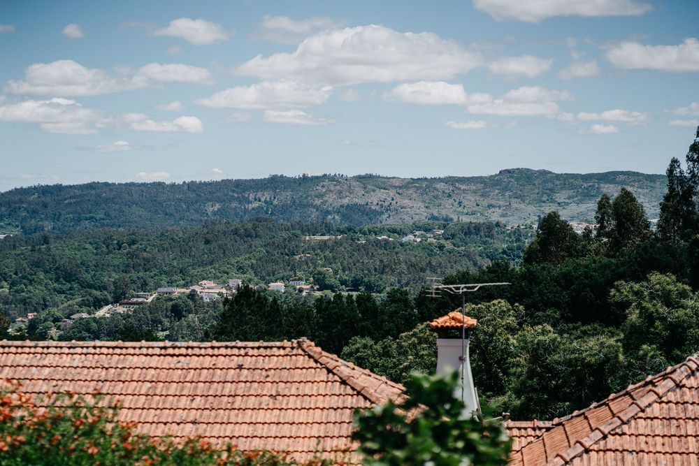 Red terracotta roof tiles in foreground with lush green hillside landscape and partly cloudy blue sky in background.