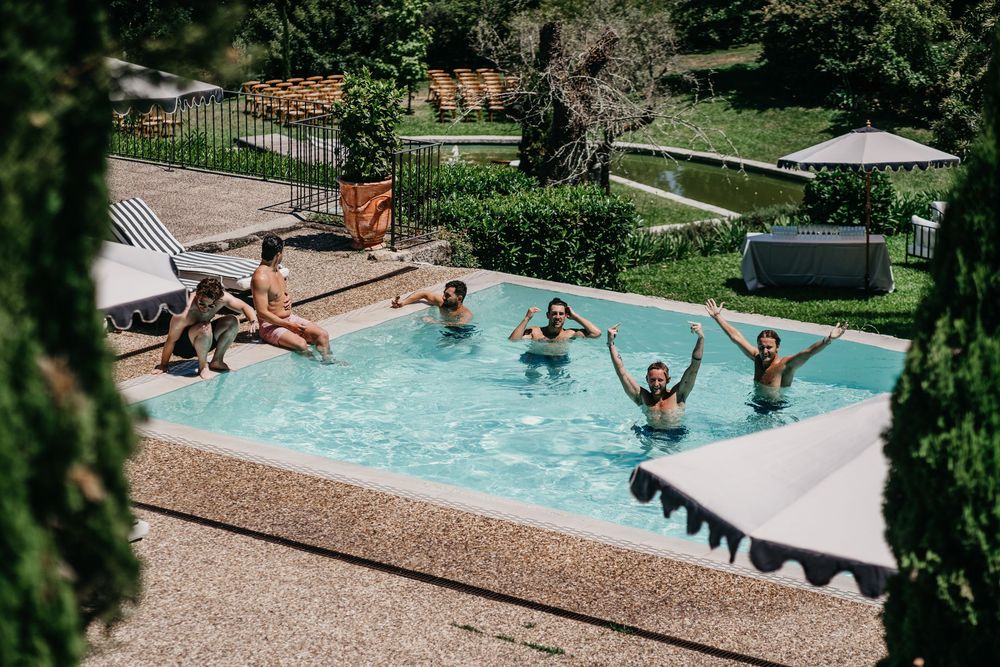 Group of friends enjoying a sunny day swimming and splashing in an outdoor pool surrounded by lush greenery.