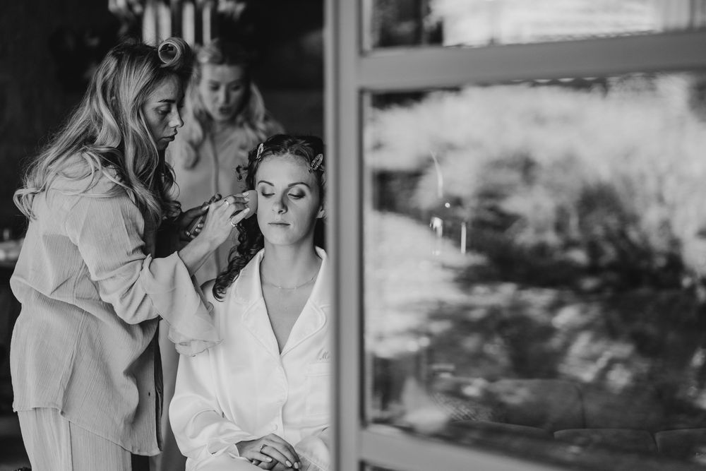 Black and white photo of a bride in a white robe getting makeup applied by a professional artist before her wedding.