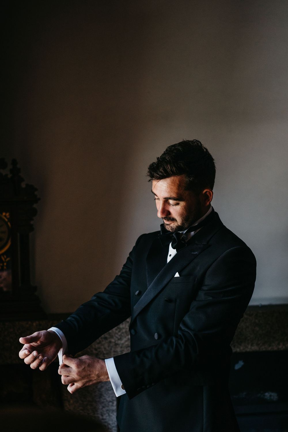 Elegant groom in black tuxedo with bow tie adjusting his cufflinks in a dimly lit room.