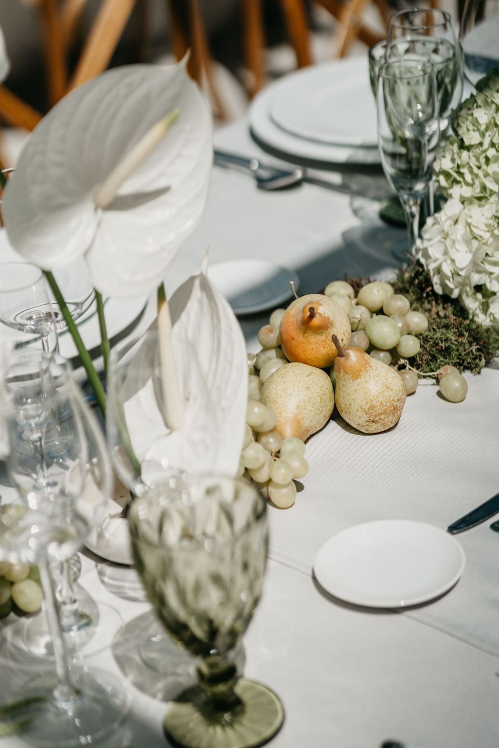 Elegant wedding table setting with white anthurium flowers, green grapes, pears, hydrangeas, and glassware on white linen.