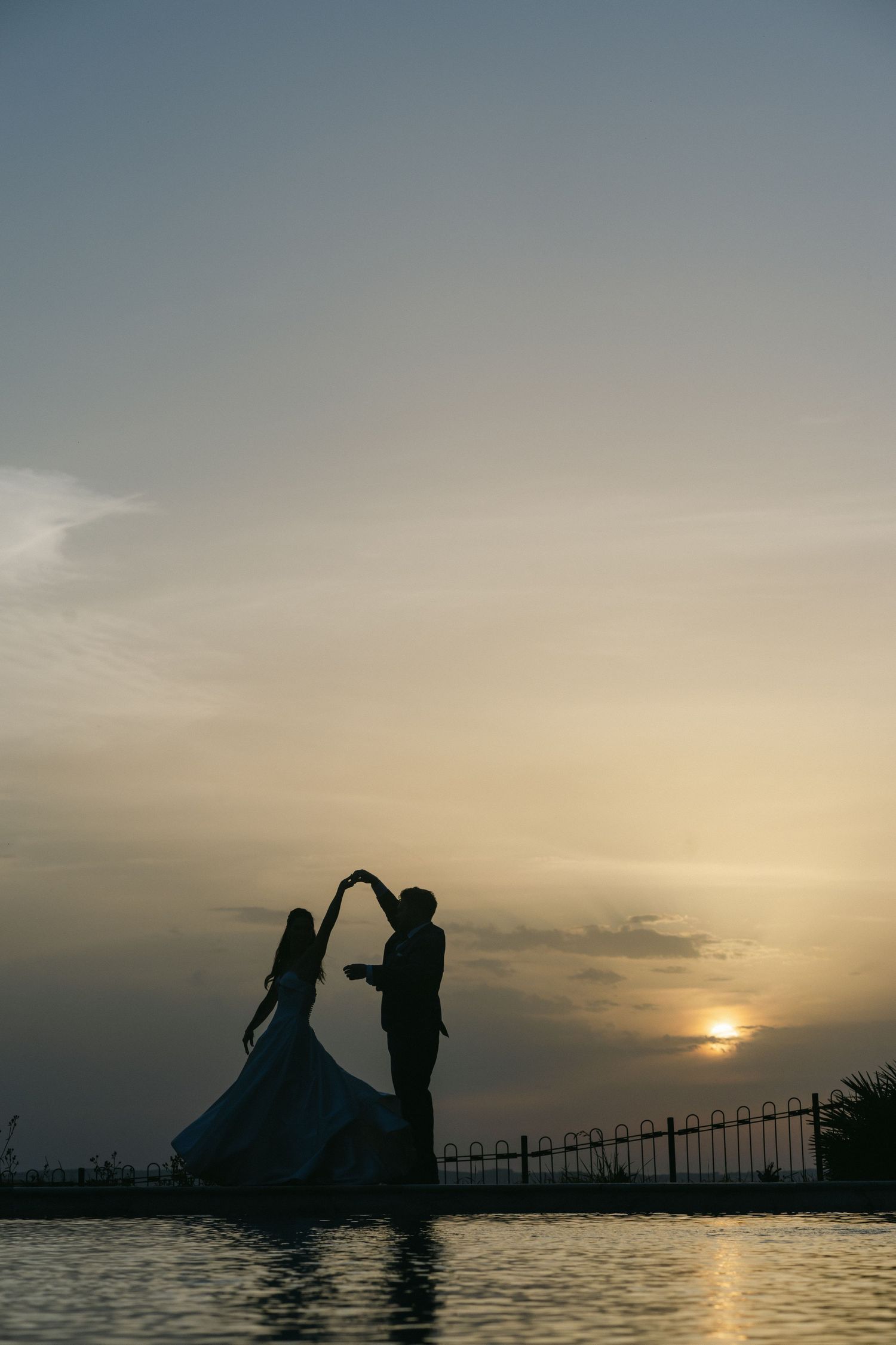 Bride and groom celebrate at sunset on a wooden deck near a pool, bride twirling in white gown, groom in blue suit.