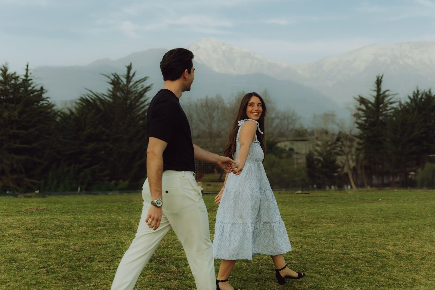 Couple walking hand in hand on a green field with snow-capped mountains in the background.