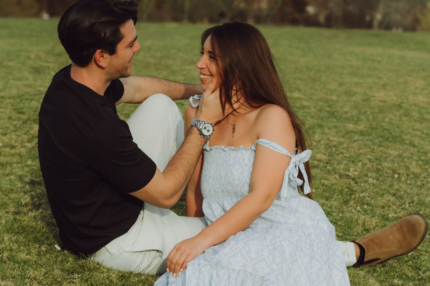 Couple sitting on grass smiling at each other during golden hour outdoor shoot.