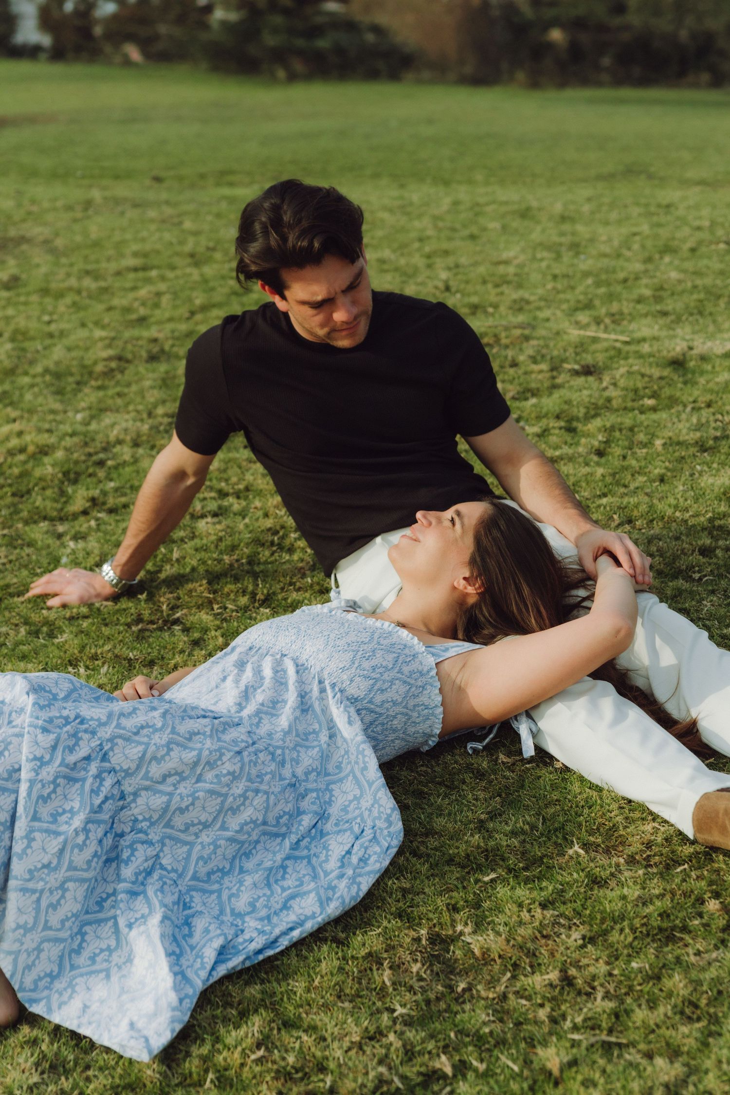 Couple relaxing on grass, woman lying down, man gazing at her lovingly outdoors.