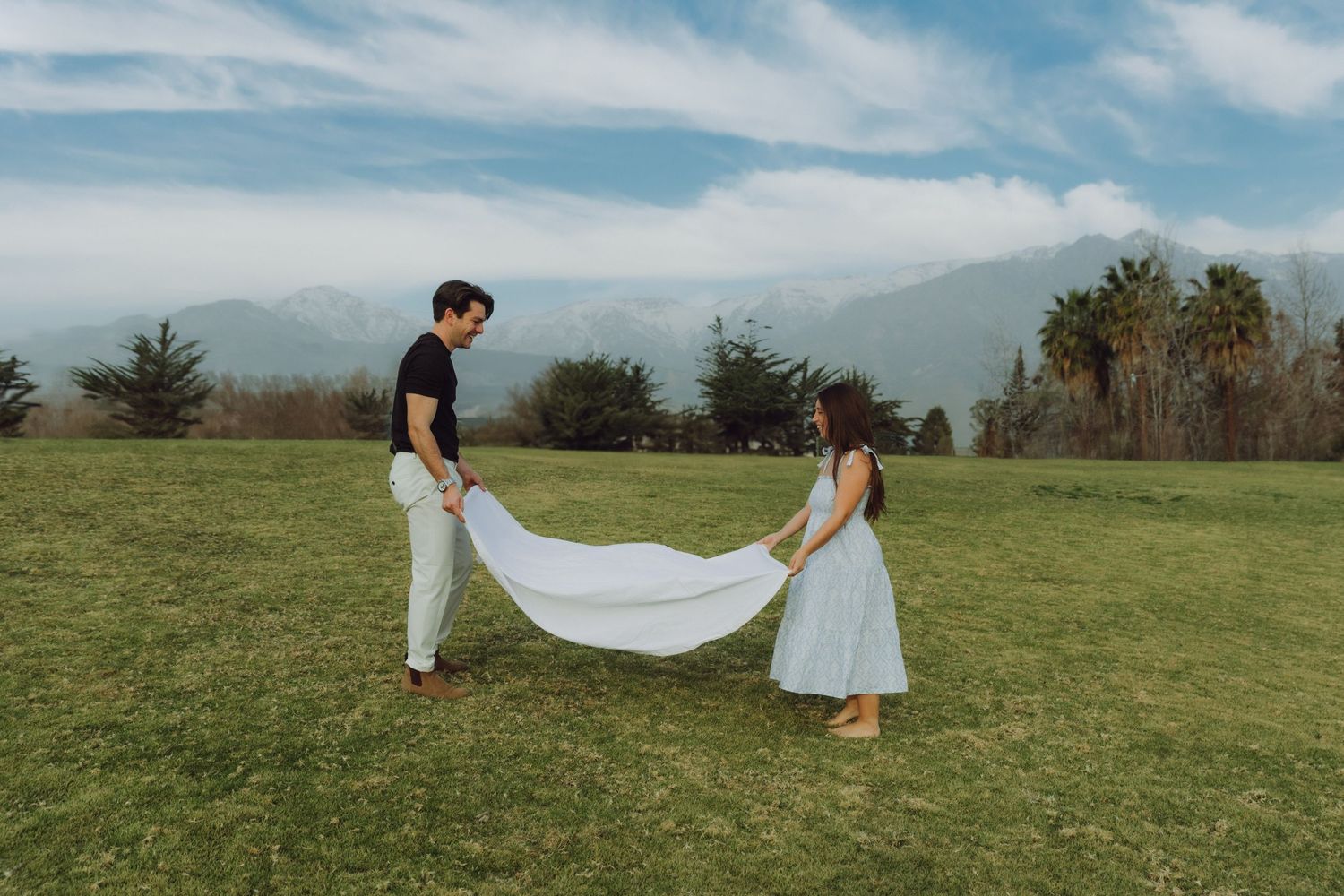 Couple spreading white blanket on green field with mountains and cloudy sky behind.