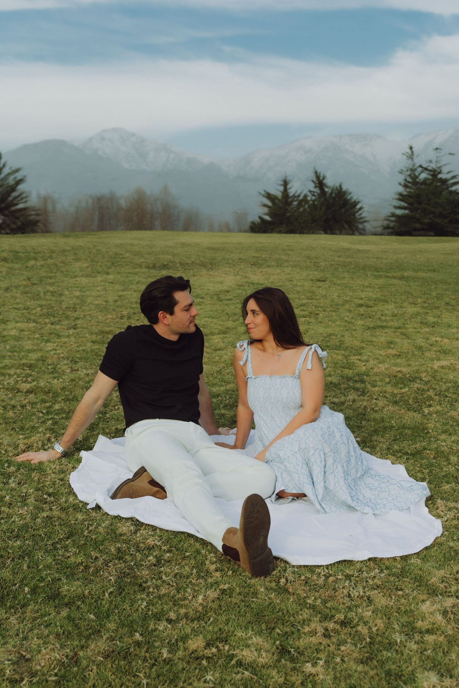 Couple sitting on white blanket on grass with snow-capped mountain range backdrop.