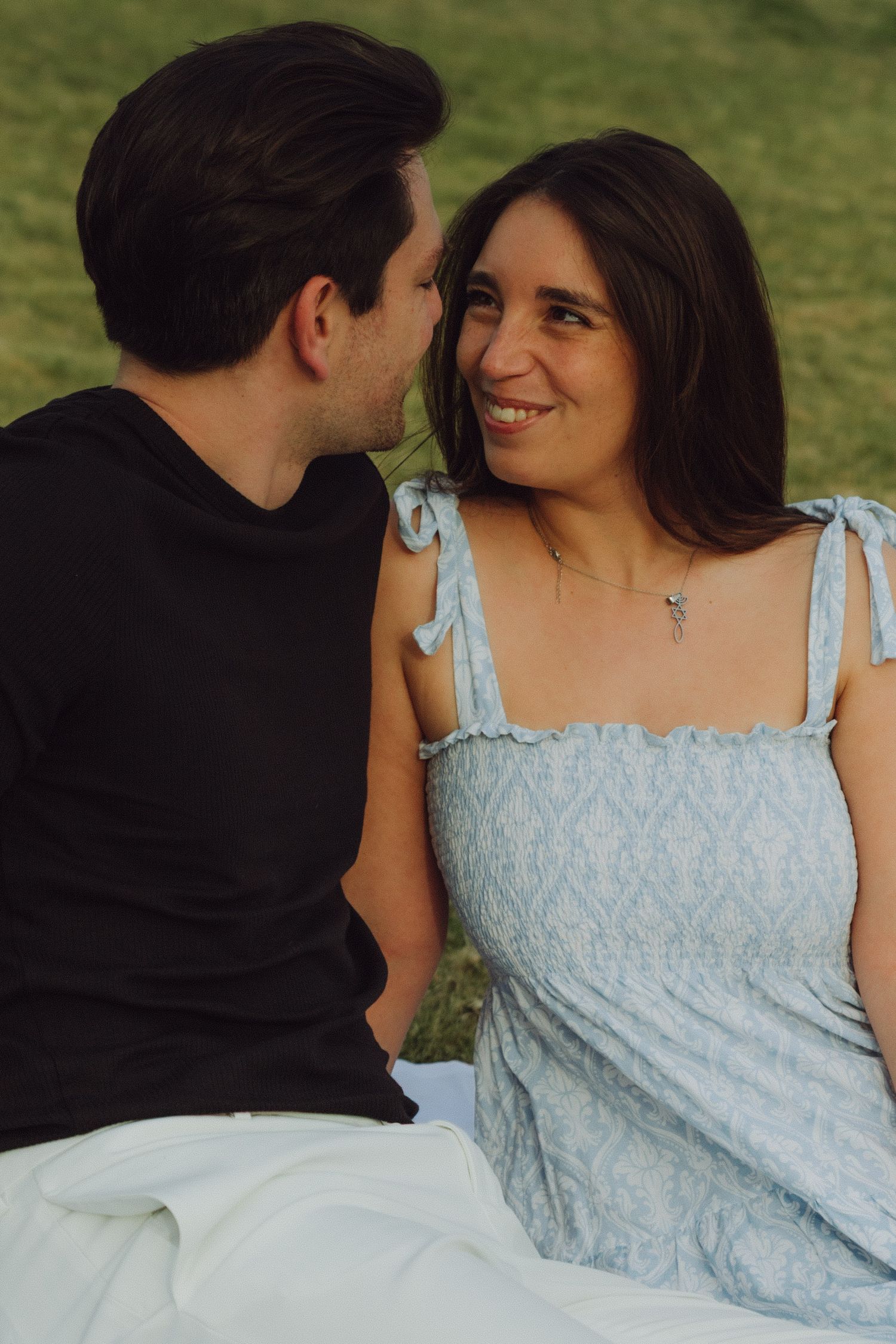 A couple sitting on grass, smiling at each other, woman in light blue dress and man in black shirt.
