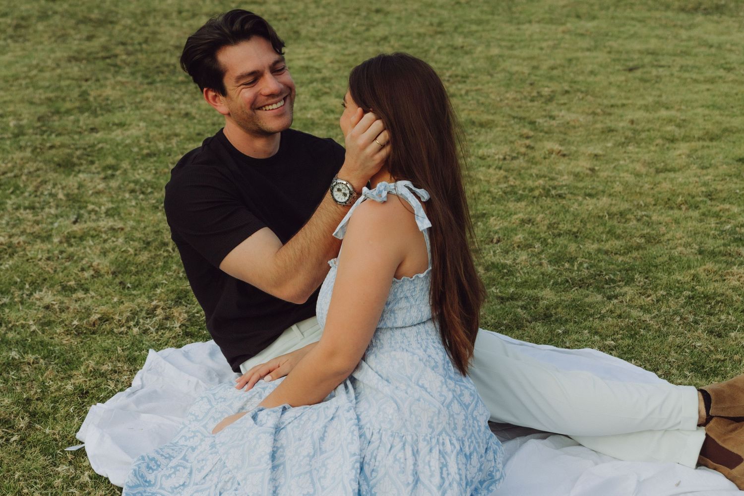 Happy couple sitting on white blanket in park, man touching womans hair, both smiling joyfully.