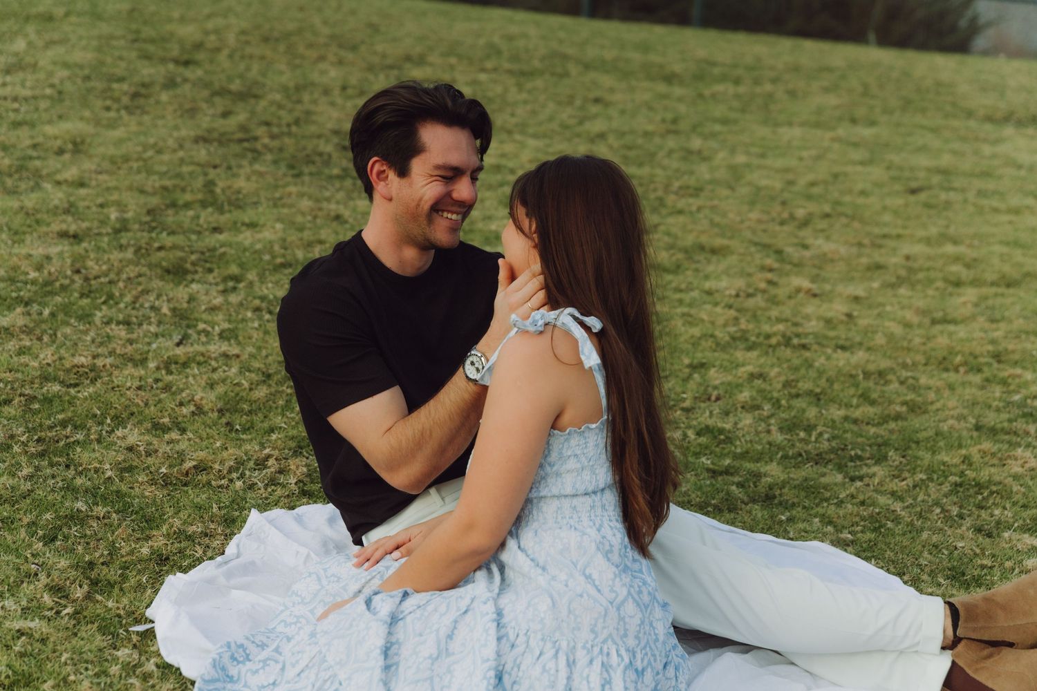 Laughing couple sitting on grass blanket outdoors, man in black shirt and woman in blue dress.