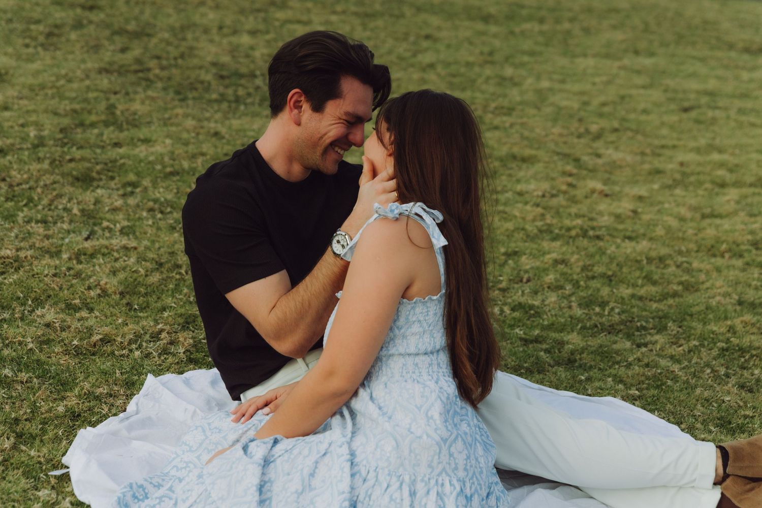 Romantic couple leaning close together on grass, woman in light blue dress, man in black shirt.