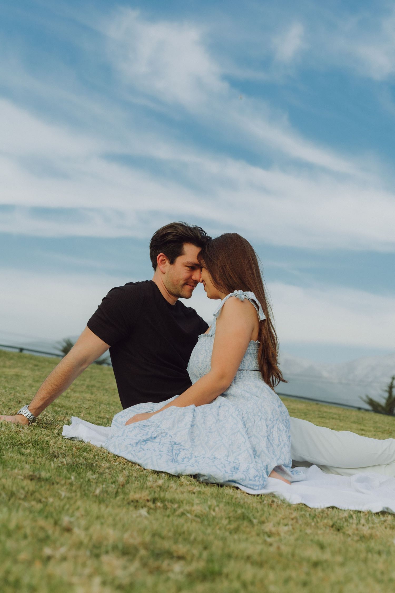 Couple sitting on grass blanket touching foreheads under blue sky with mountains in background.