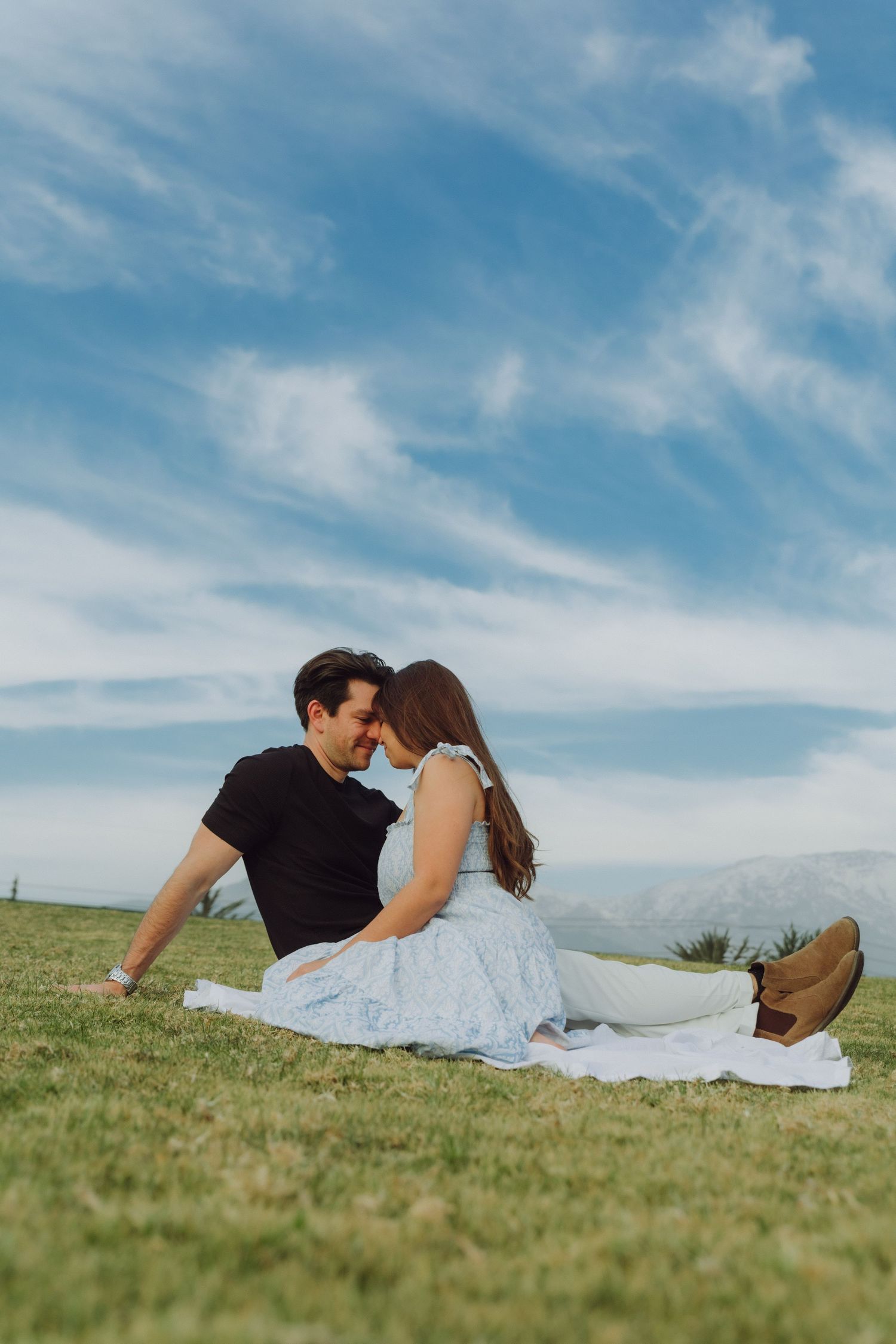 Couple sitting on white blanket on green hill, leaning toward each other under a cloudy blue sky.