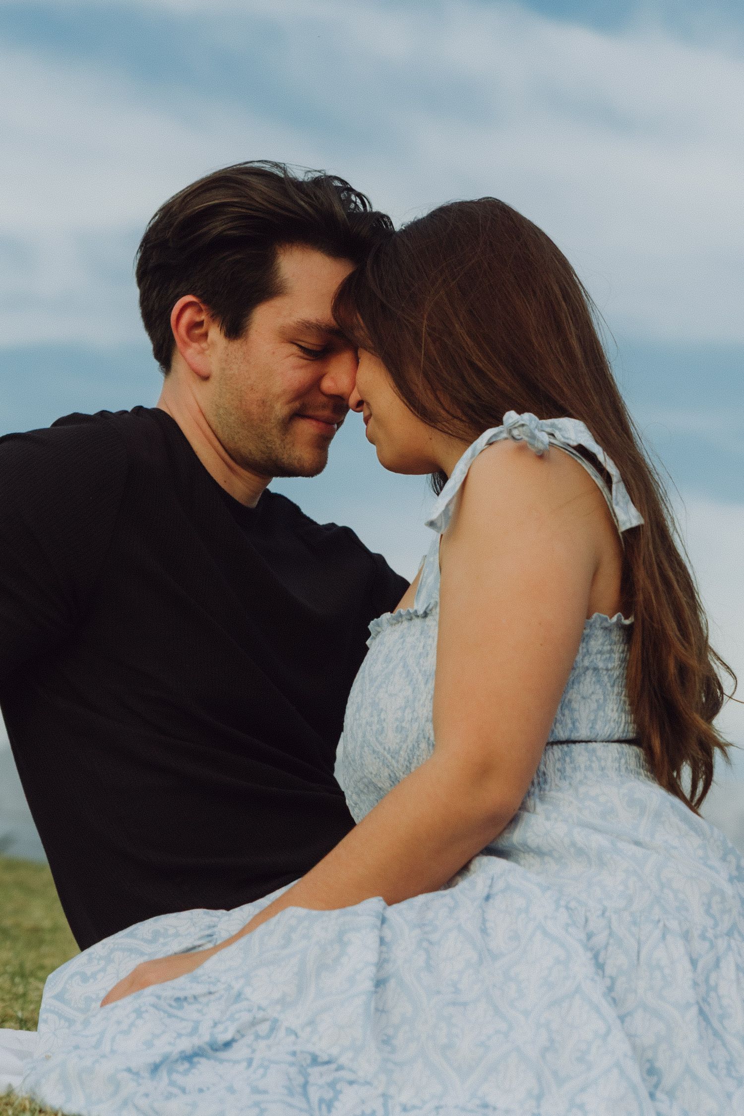 Close-up of couple touching foreheads tenderly outdoors, man in black shirt and woman in blue dress.