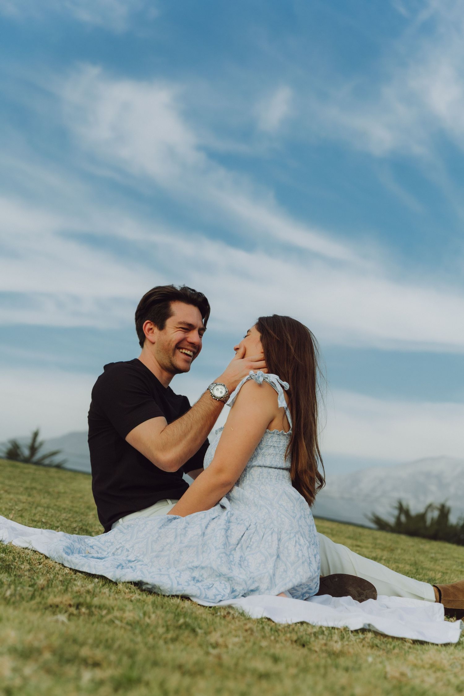 Smiling couple sitting on grass blanket outdoors, man laughing and touching womans face under blue sky.