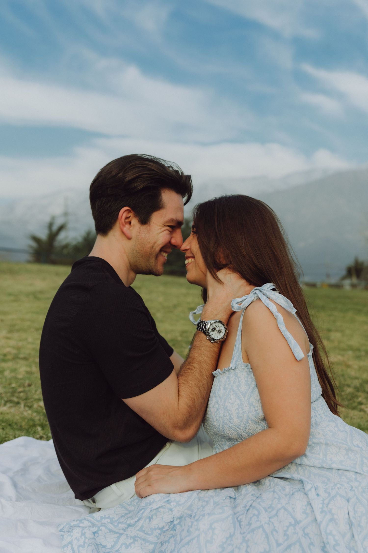 Happy couple smiling face to face on grass blanket with mountains and blue sky in background.