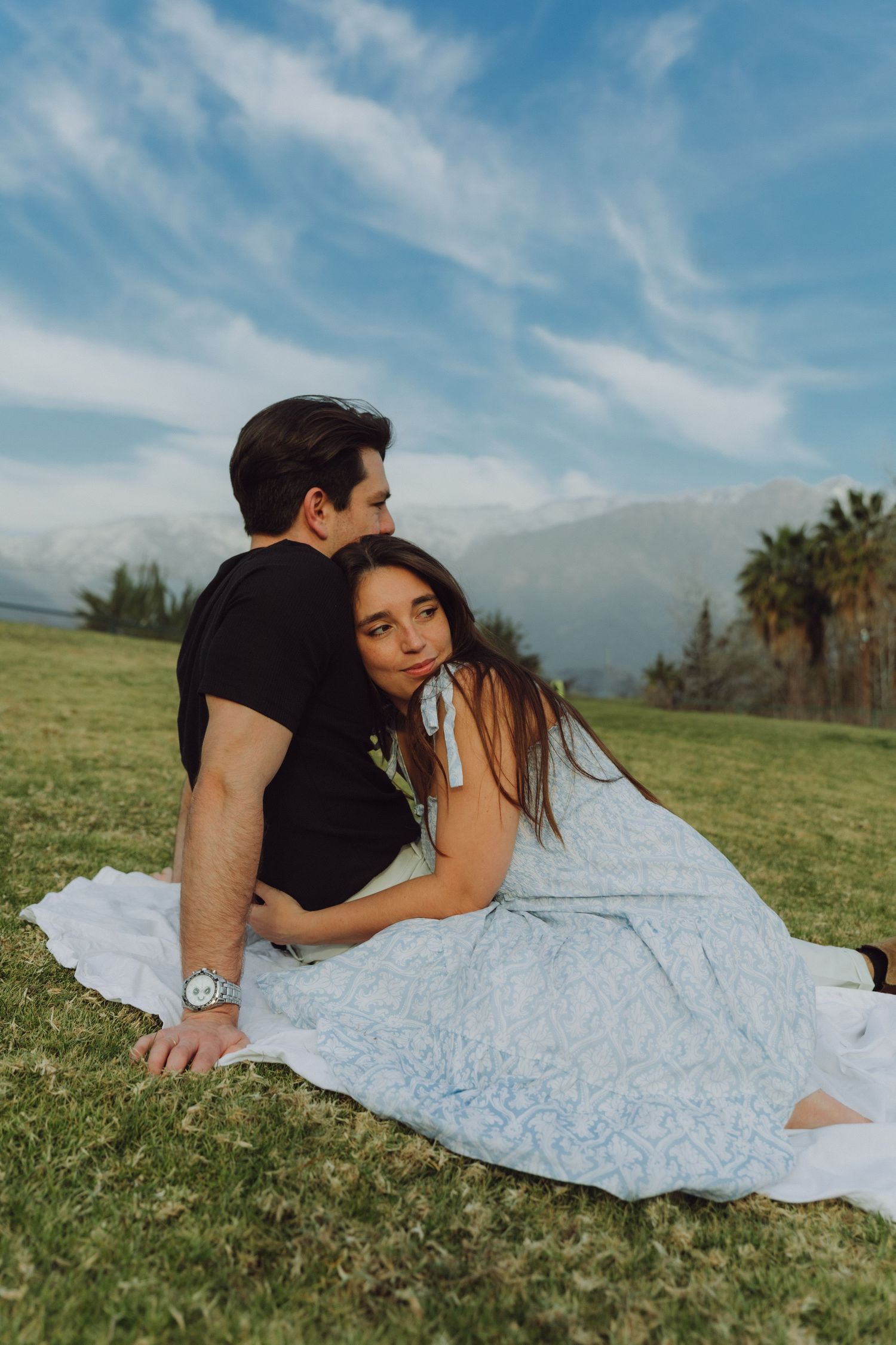 Woman resting head on mans shoulder while sitting on blanket outdoors with mountains behind them.