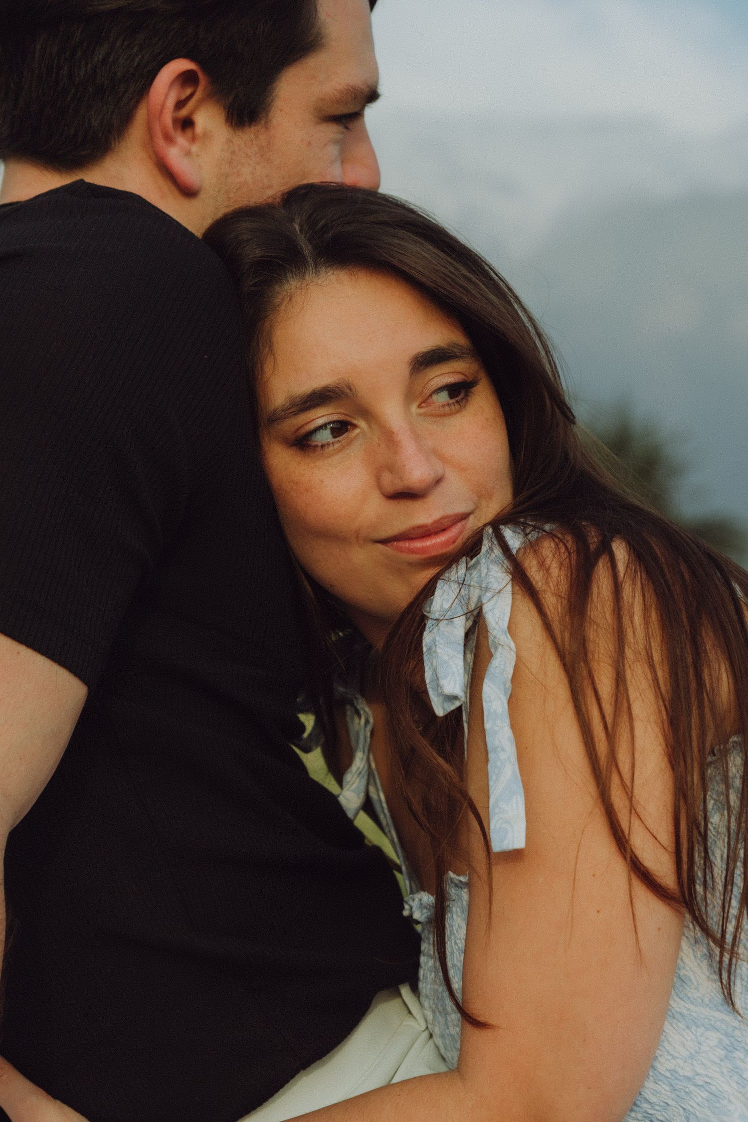 Young woman with long brown hair rests her head on a man's shoulder in a romantic outdoor embrace.