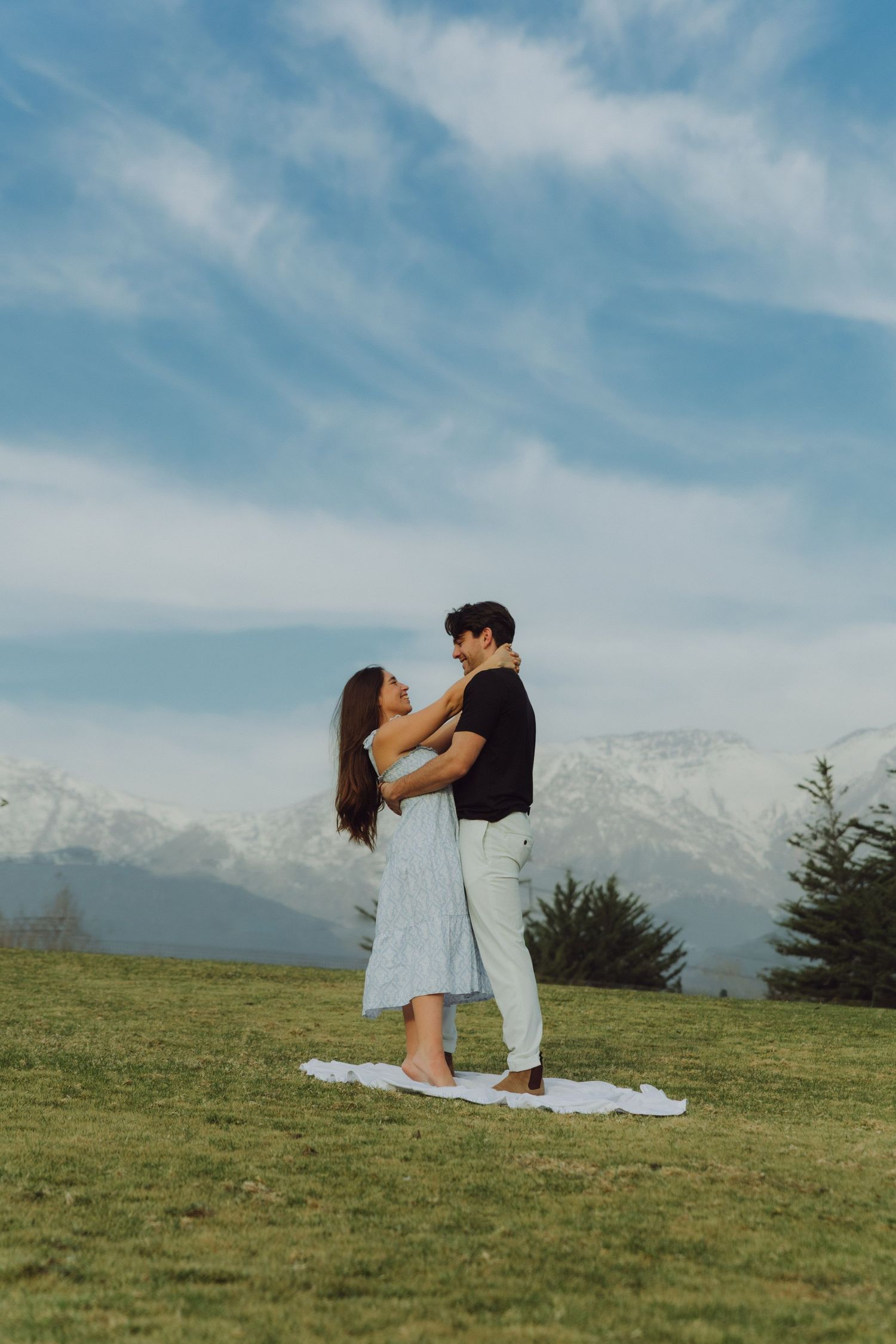 Couple dancing together on a blanket in a meadow with snow-capped mountains under a blue sky.