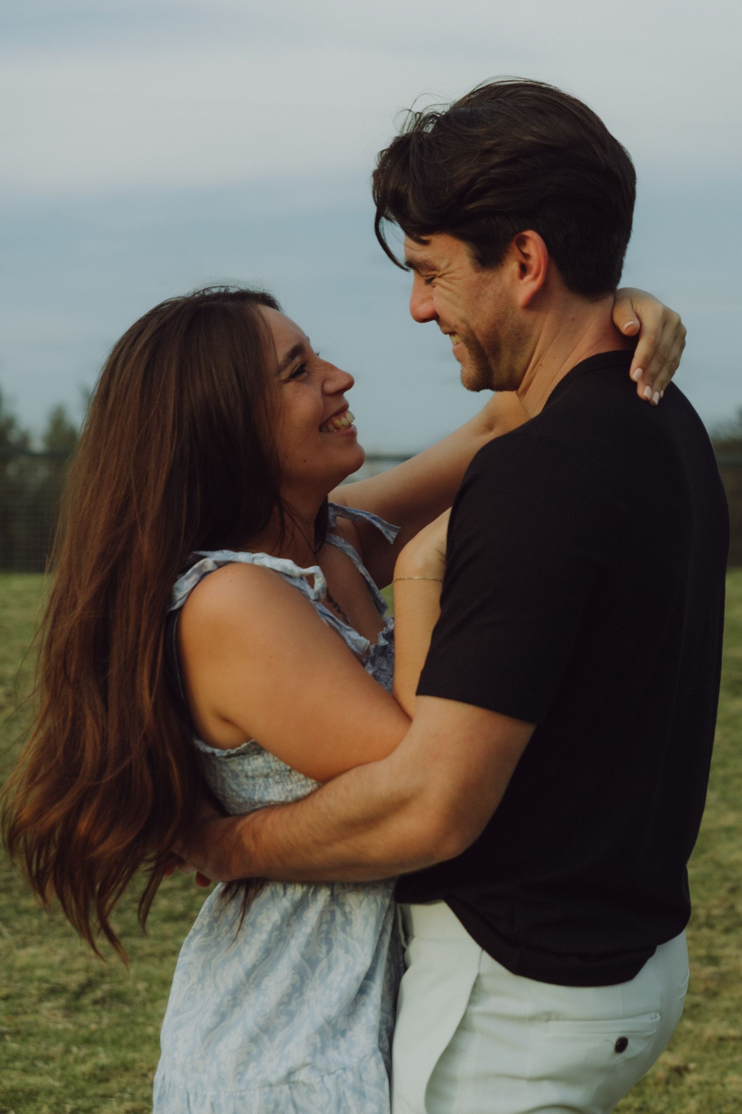 Happy couple embracing and smiling at each other in a green field during golden hour.