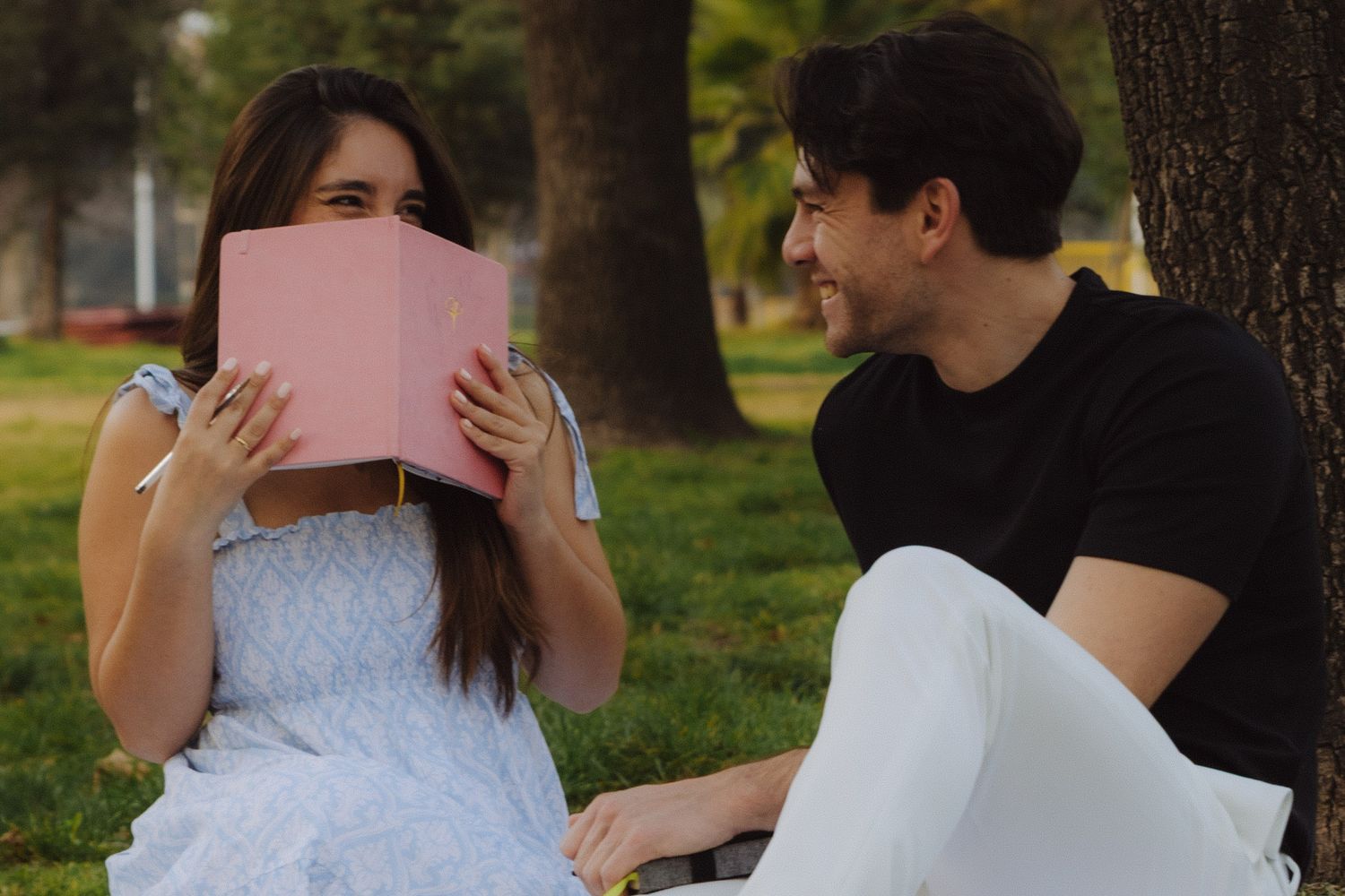 Young woman playfully hides her face behind a pink book while a man smiles at her in a park.