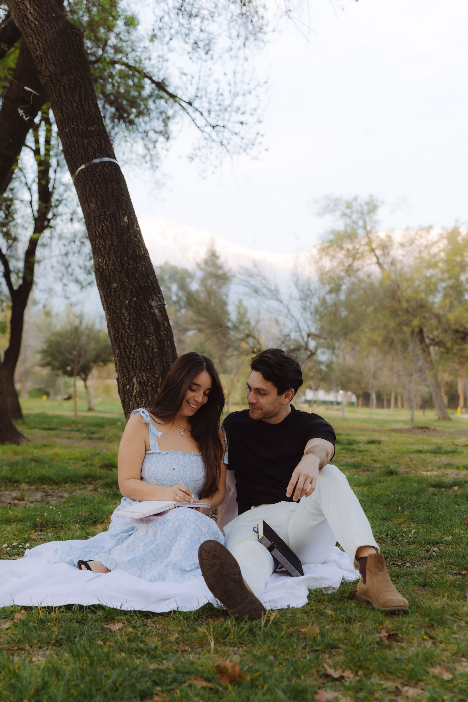Couple sitting together on a blanket leaning against a tree in a peaceful park setting.