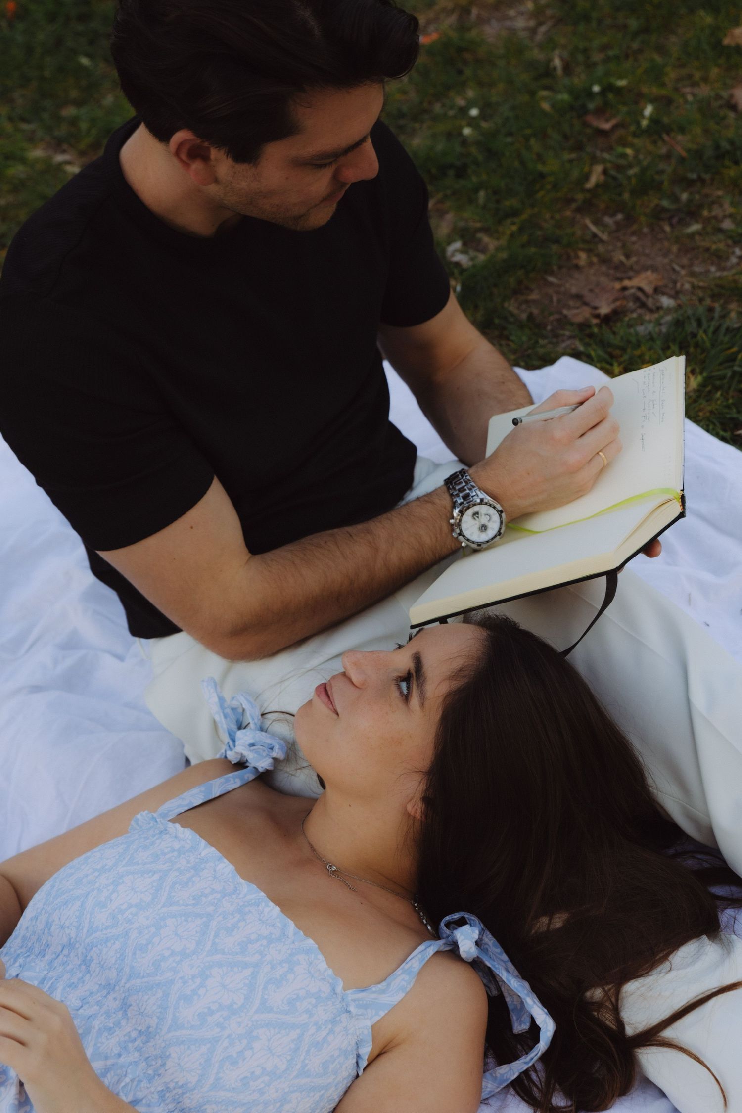 Man reading aloud from a book to a woman lying on a blanket in an outdoor setting.