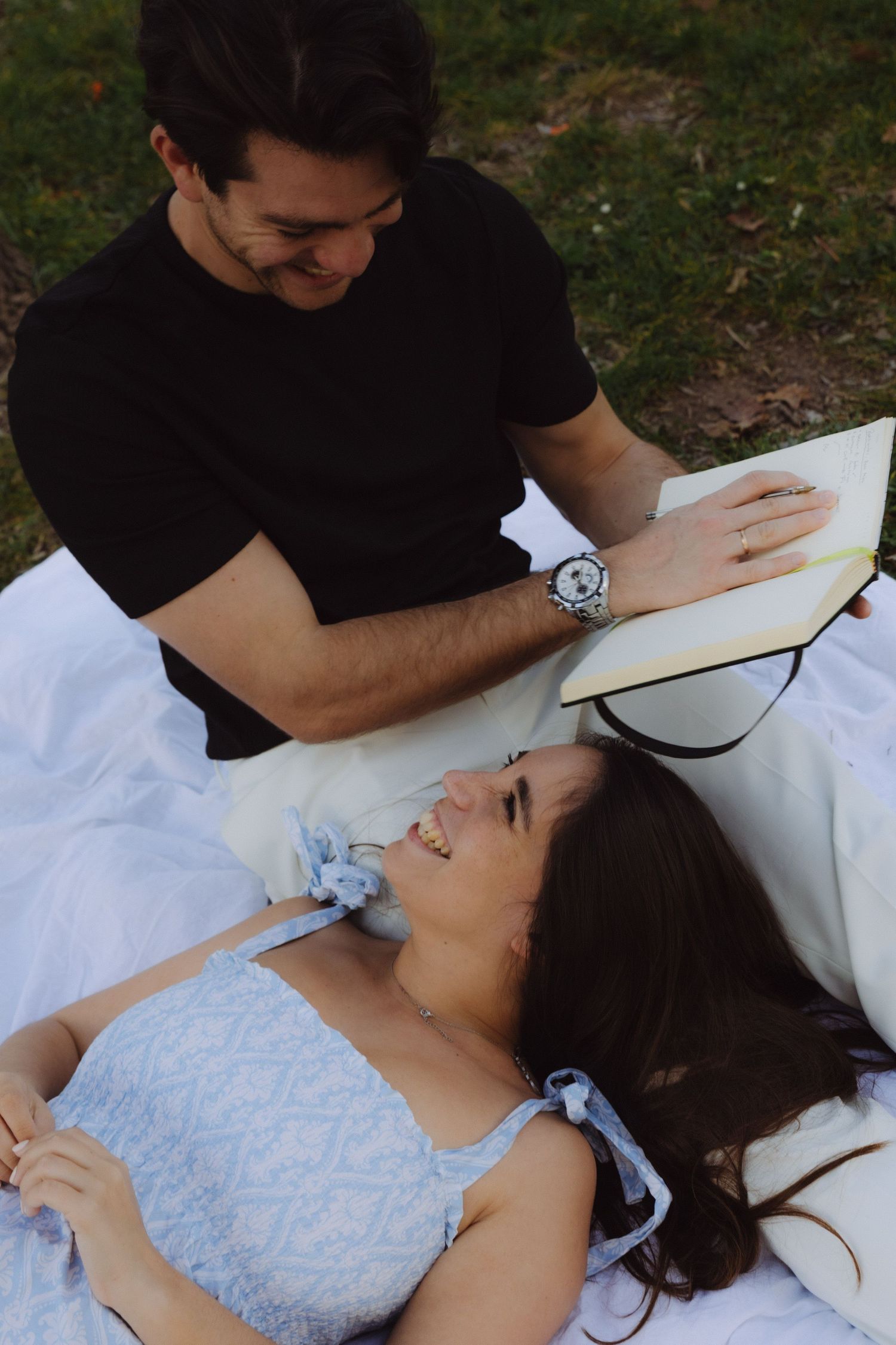 Laughing couple sharing a joyful moment on a blanket outdoors, man holding an open book.