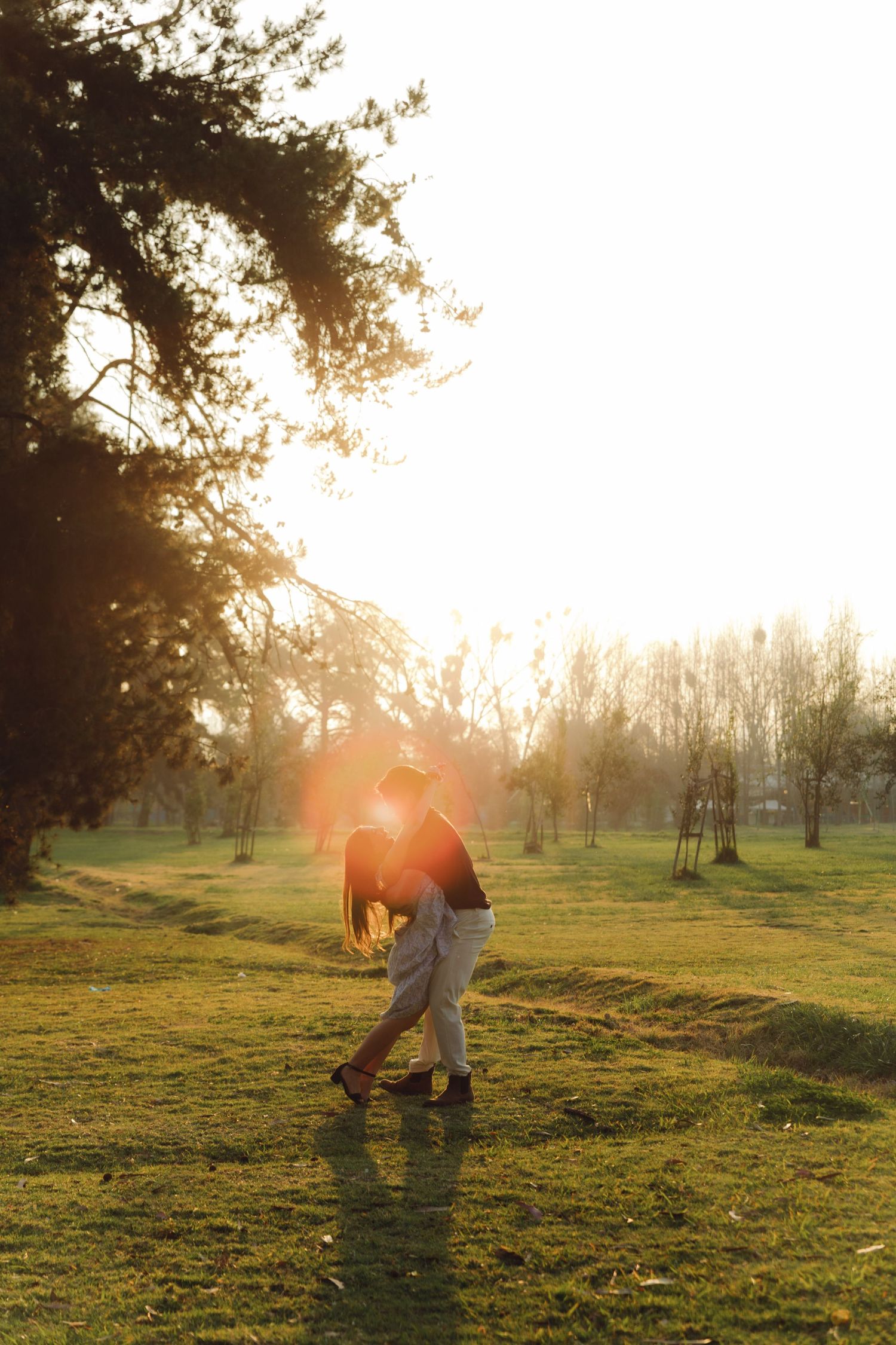Couple embracing in golden sunset light in a lush green park with tall trees.