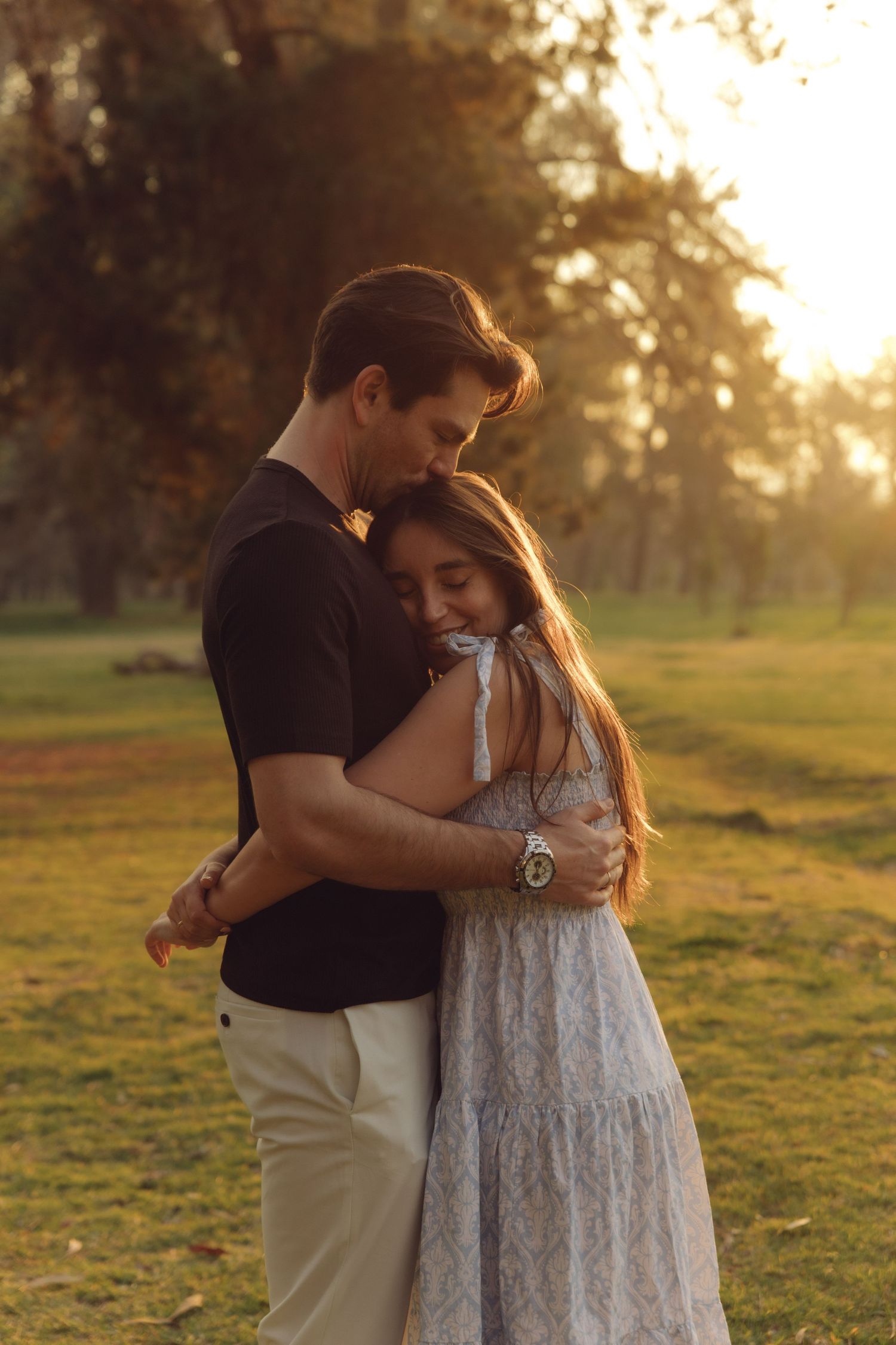 Couple embracing tenderly in a sunlit park at golden hour, man in black shirt hugging woman in white dress.