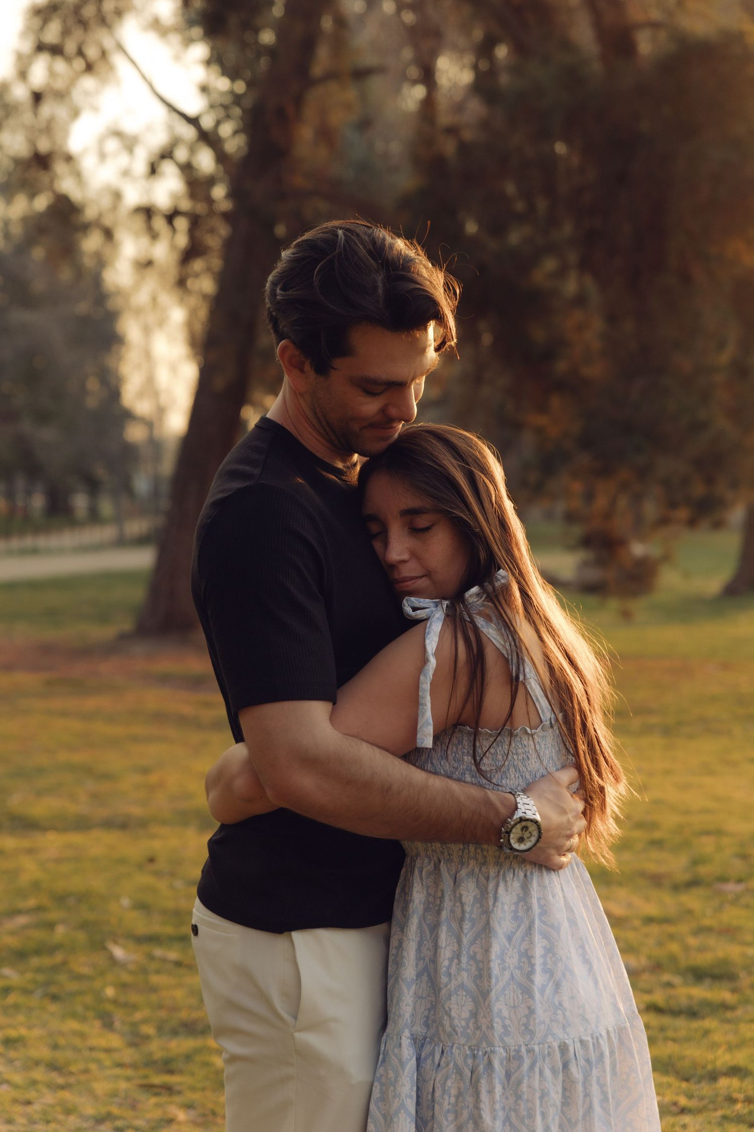 Romantic couple sharing a close embrace outdoors at sunset, surrounded by trees in a peaceful park setting.