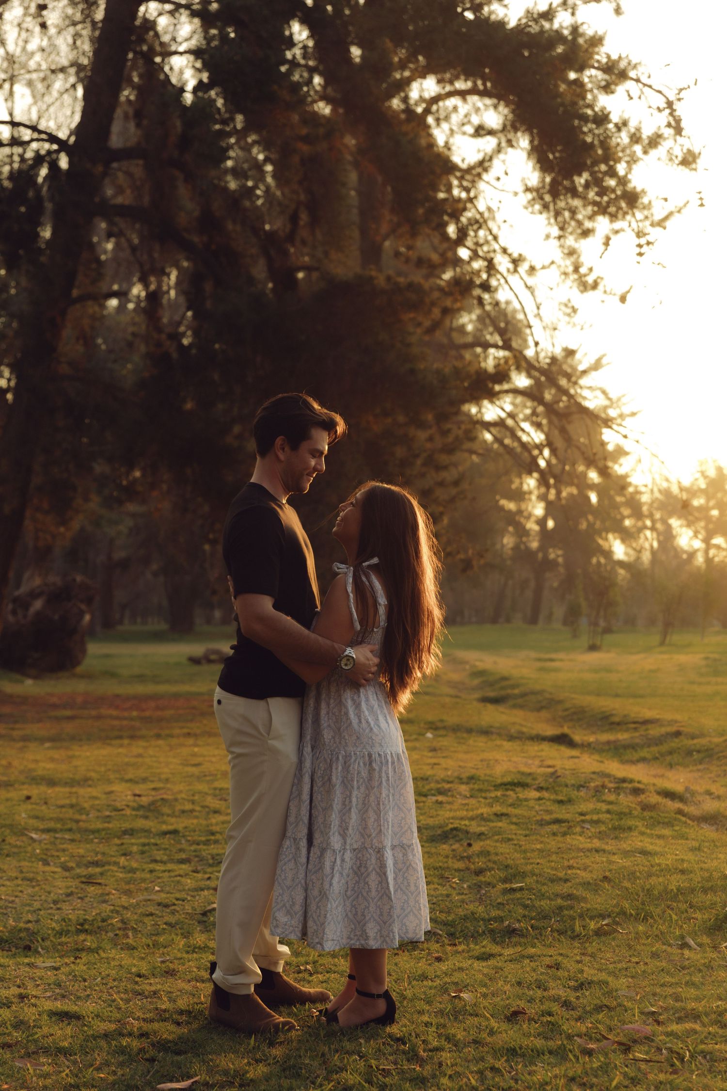 Young couple standing face to face holding hands in a golden sunset park, sharing an intimate moment.