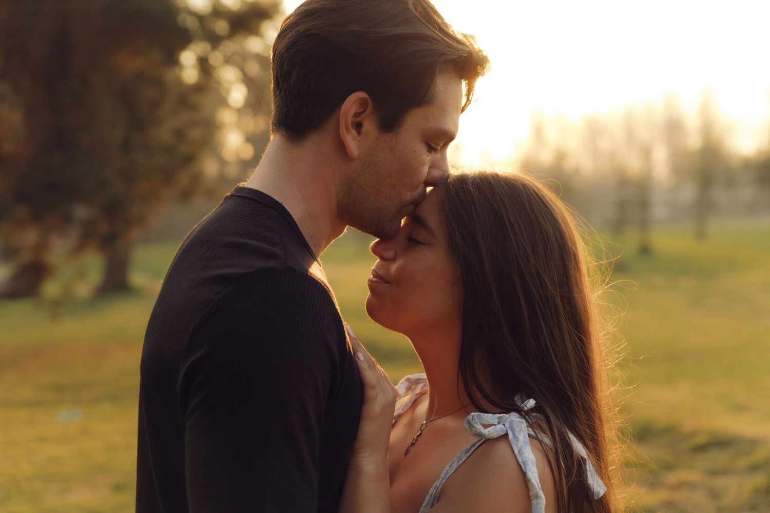 Couple leaning in for a kiss at sunset in a park, bathed in warm golden light among green trees.