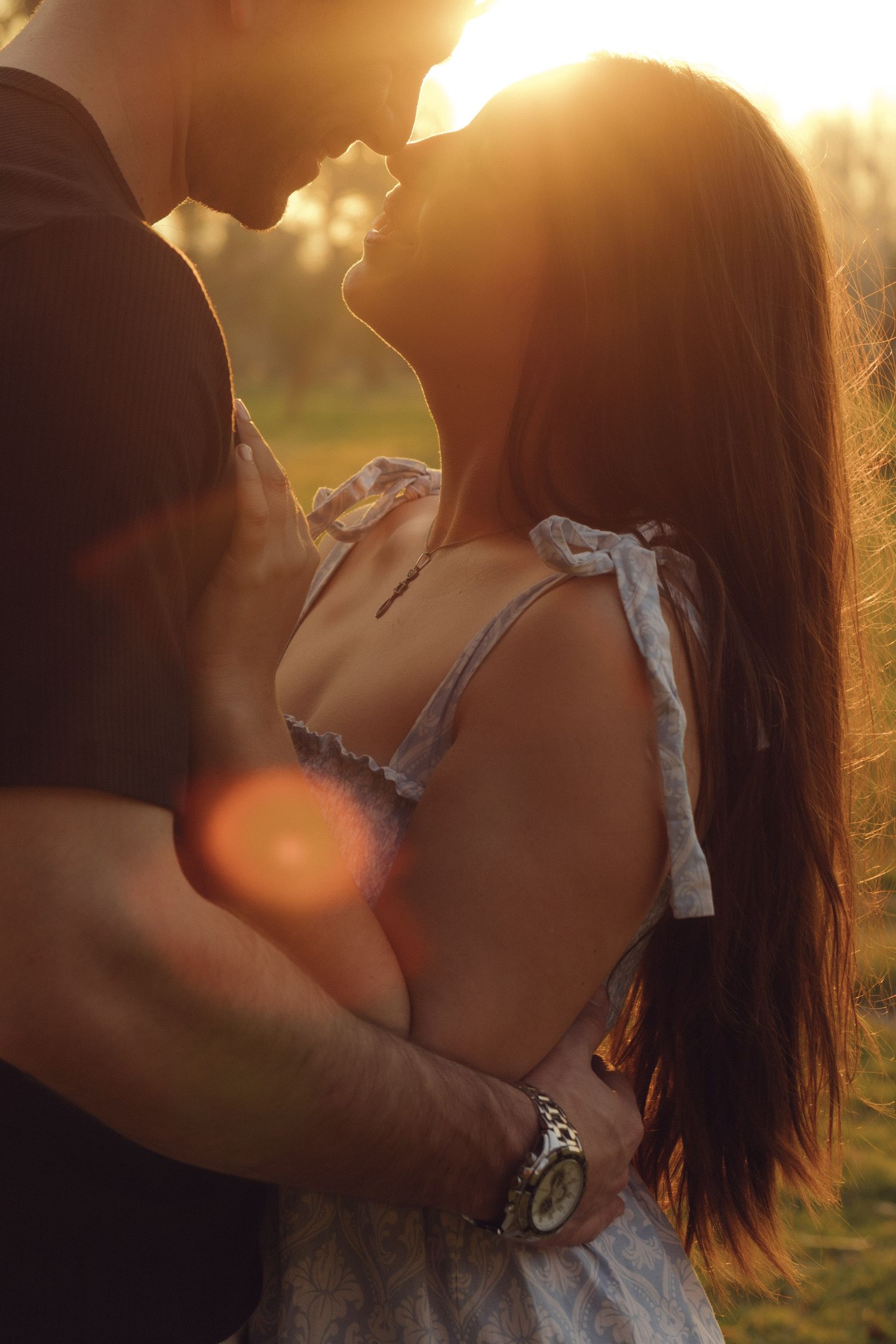Couple embracing and almost kissing in a sun-flared golden hour outdoor setting, woman in a light dress.