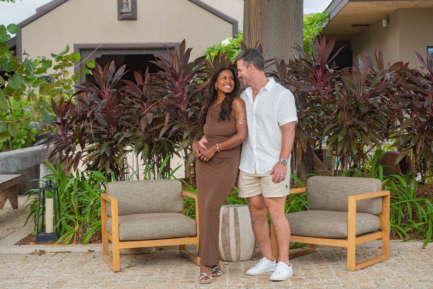 Couple posing outdoors near tropical plants, woman in brown dress, man in white shirt and shorts, patio furniture behind them.