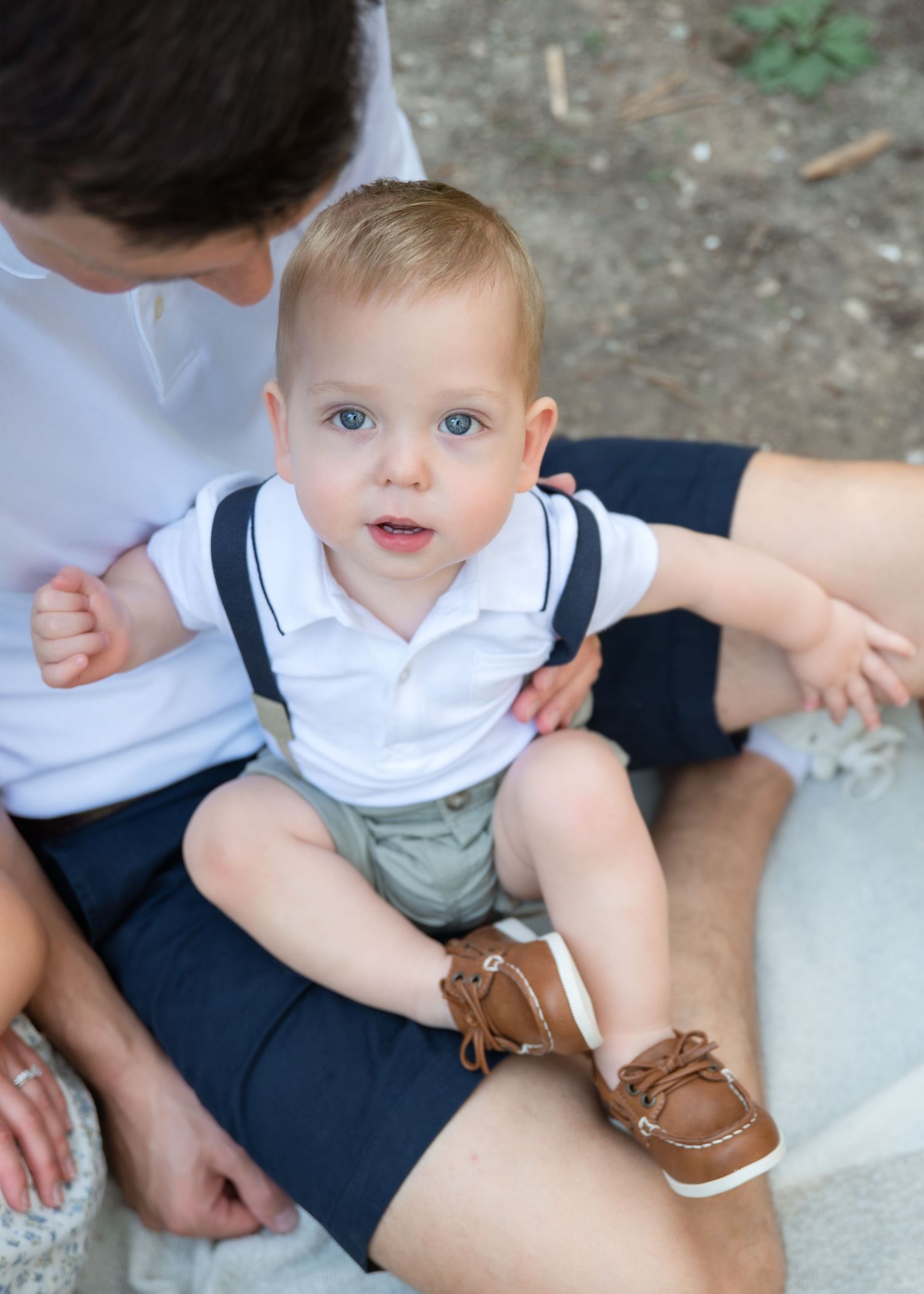 Adorable blue-eyed baby boy in white polo, suspenders, and brown boat shoes sitting on father's lap outdoors.