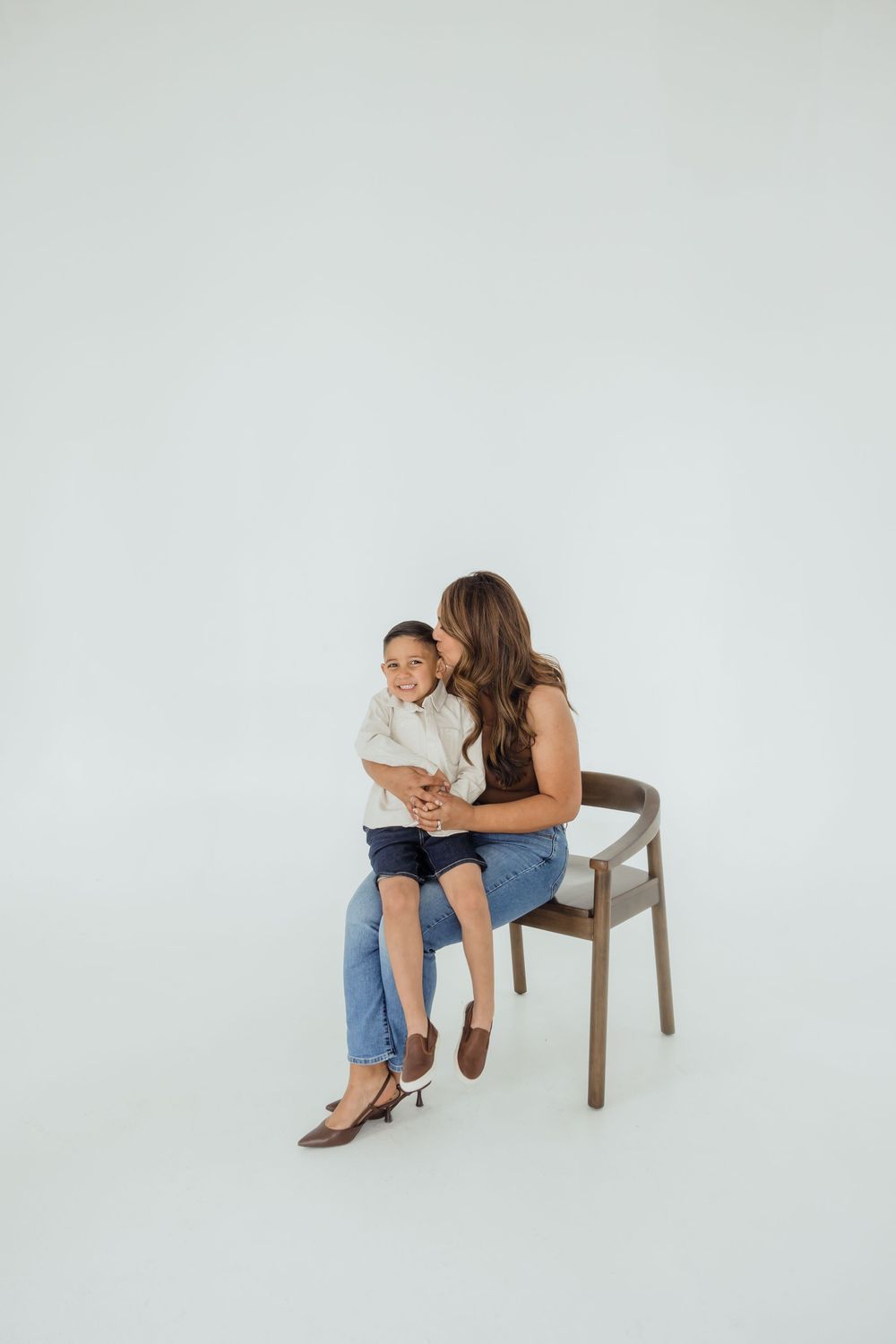 A woman sits on a wooden chair hugging a smiling child in a bright studio setting, both wearing casual blue jeans.