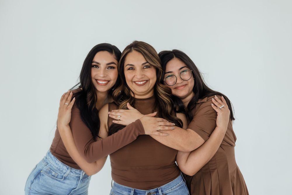 Three women in brown tops smiling and hugging each other against a light gray background.