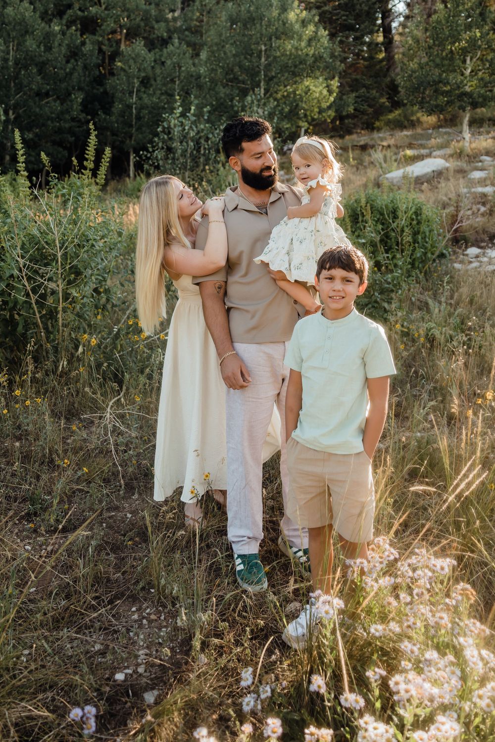A happy family of four poses outdoors in a sunlit meadow with trees, parents holding a toddler girl and a smiling boy in front.