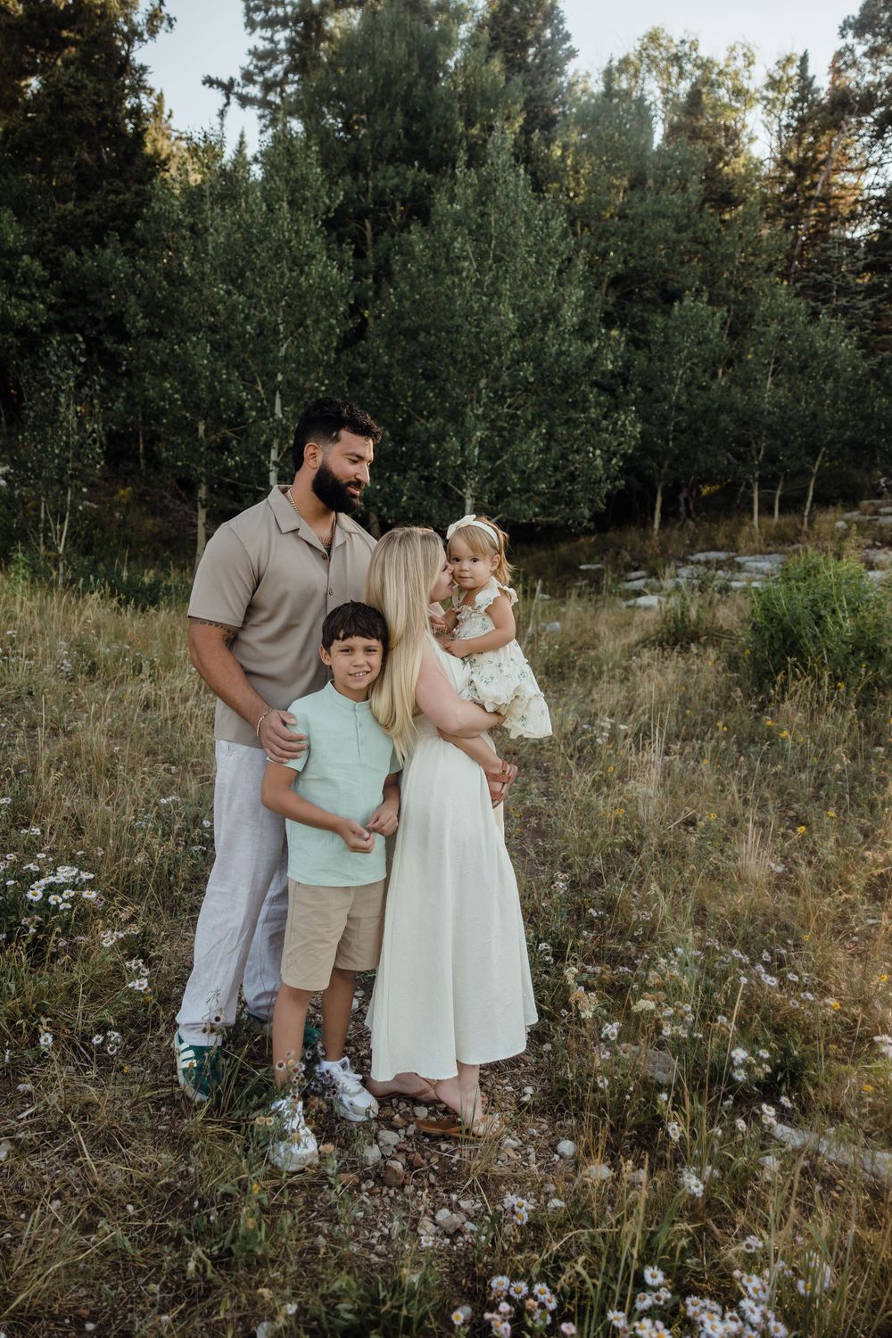 A bearded father and blonde mother hold their young children in a sunlit meadow surrounded by green trees.