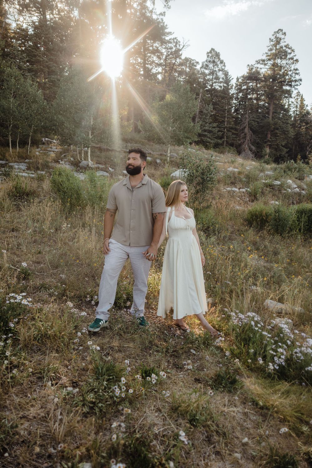 Couple holding hands in a sunlit forest meadow, man in gray shirt and woman in white dress, with pine trees backdrop.