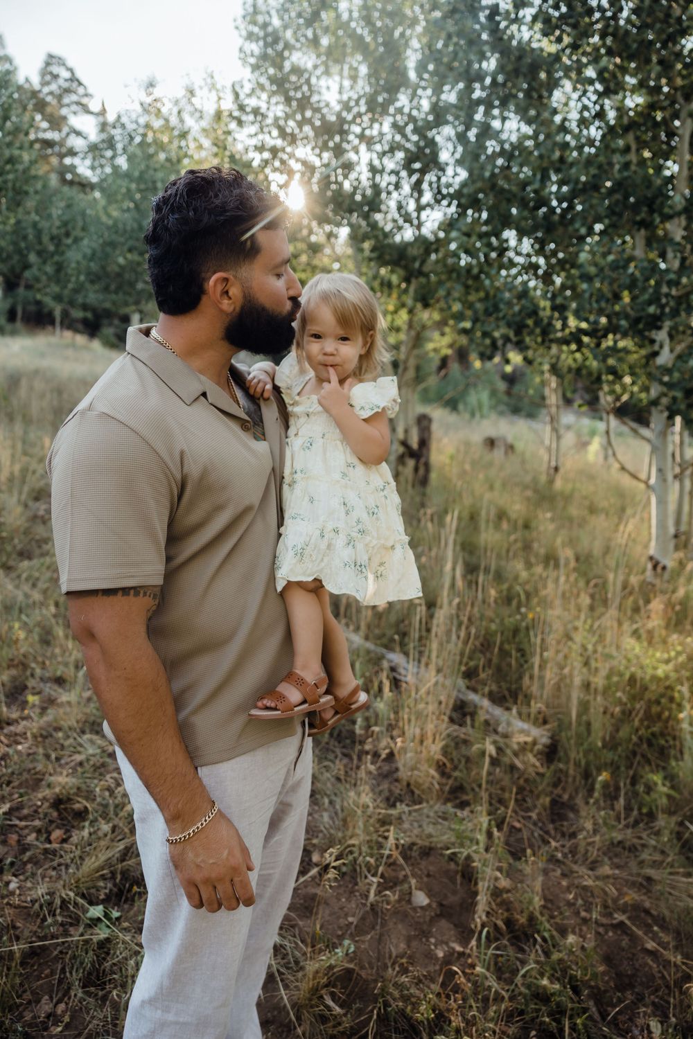 A bearded man in a tan polo holds a toddler girl in a white floral dress outdoors in a sunlit forest setting.