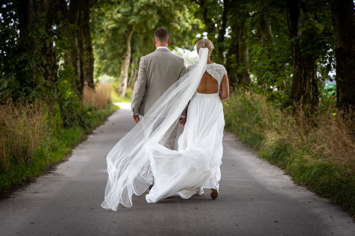 Bride and groom walking away down a tree-lined path, wedding dress veil flowing in the breeze.
