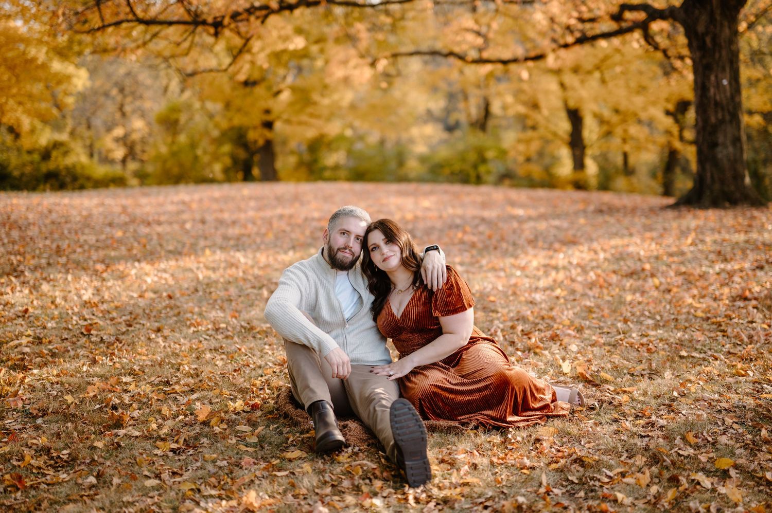 Couple sitting together on fall leaves in a park, surrounded by golden autumn trees.