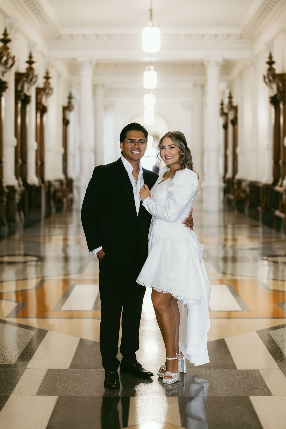 Couple in wedding attire posing in elegant marble hallway, groom in black suit, bride in white mini dress.